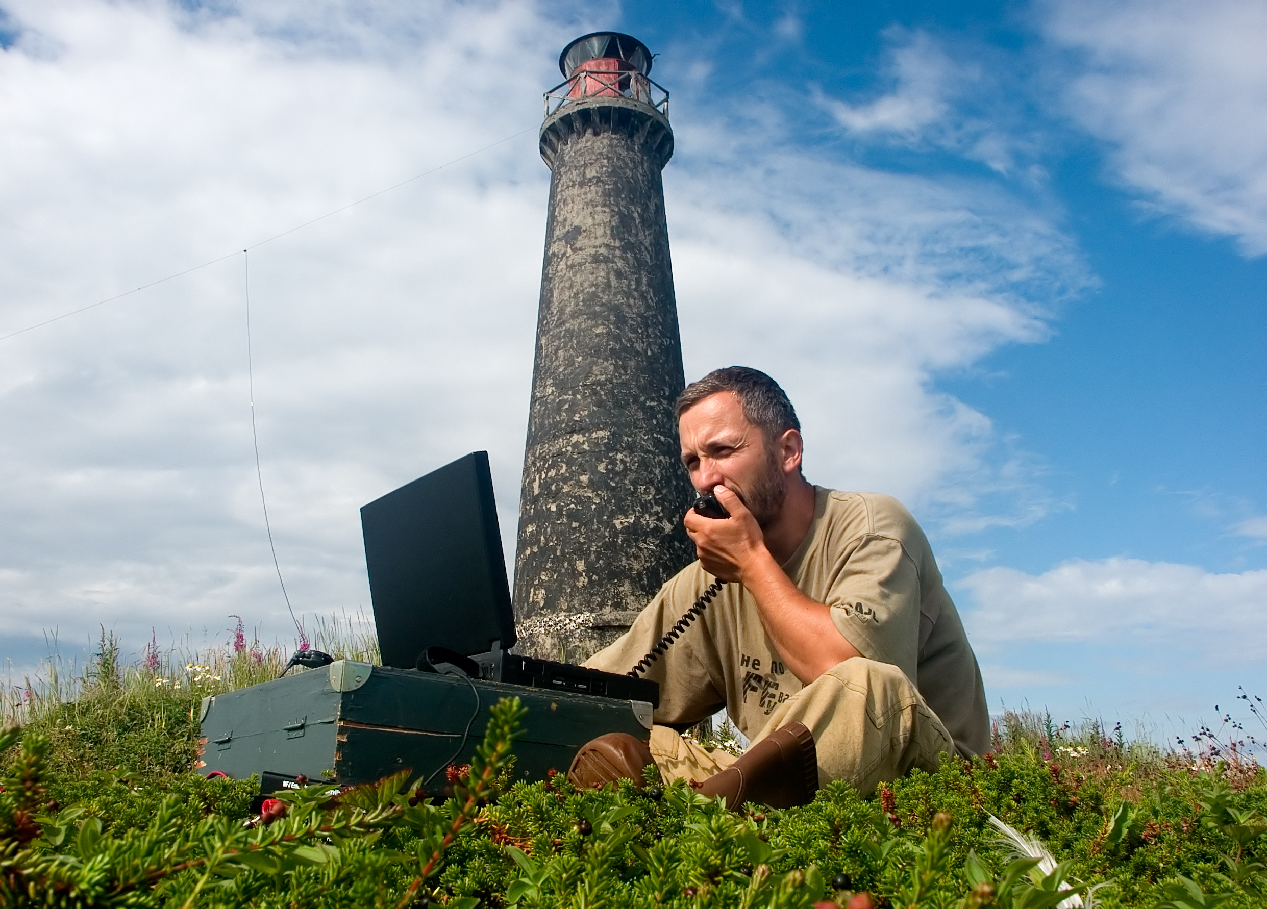 white middle-aged man sitting on a mountainside on a sunny day, using a CB radio